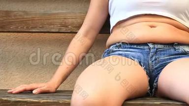 穿短裤和短T恤的胖女人坐在长凳上。 从衬衫下面可以看到腹部脂肪的褶皱